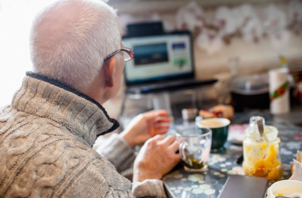 A senior man wearing a sweater sits alone at a table facing away from the camera watching TV with a cup of tea.