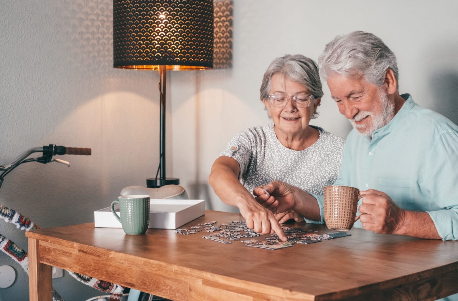 A smiling elderly couple are working on a puzzle together while sitting at a table and drinking tea