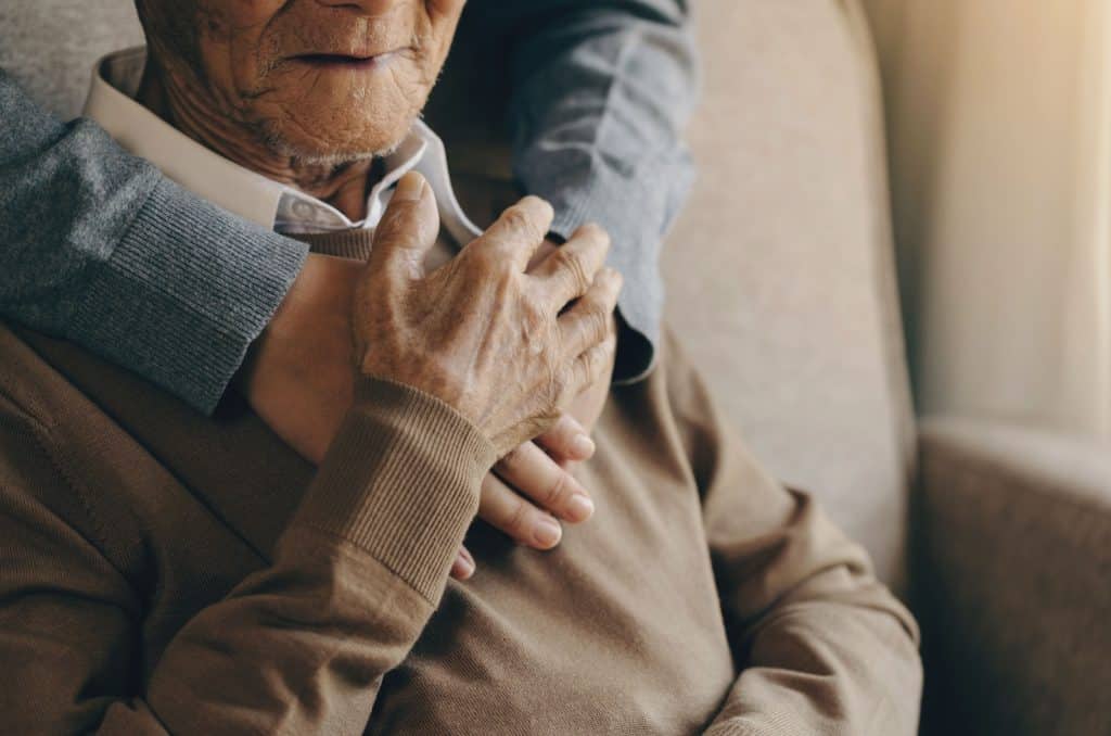 A close-up image of an older adult sitting with their right hand clasped over a caregiver's hands on their chest.