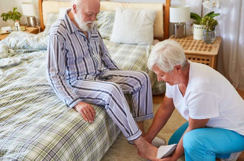 A caregiver putting slippers on an older adult's feet while they are sitting on a bed in their pajamas.