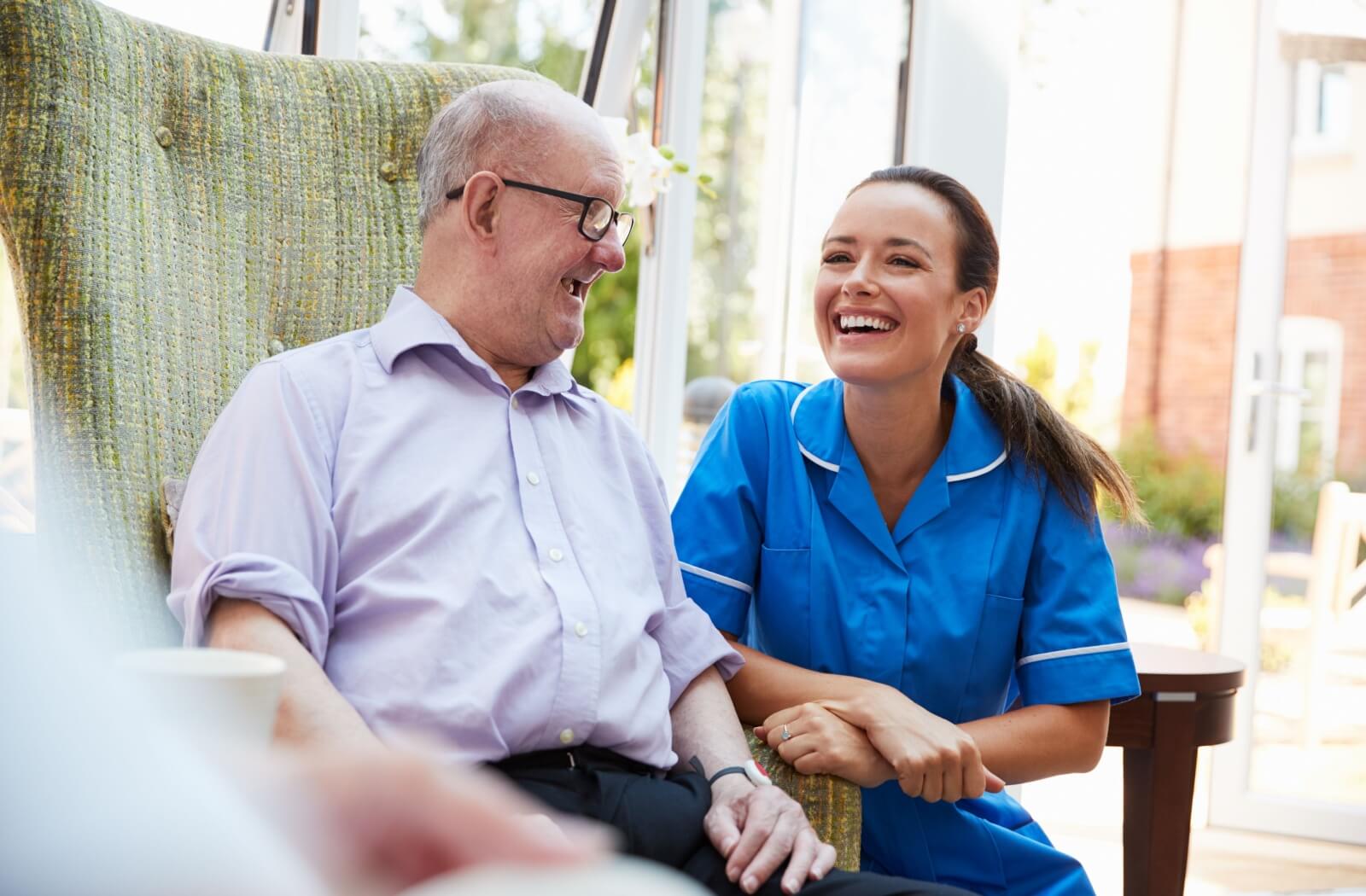 A senior at a memory care community sitting in a chair laughing while their memory care nurse sits smiling beside them.
