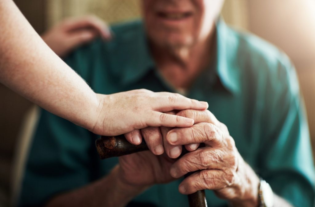 A senior adult holds onto their cane as a caregiver rests their hand on top of theirs.